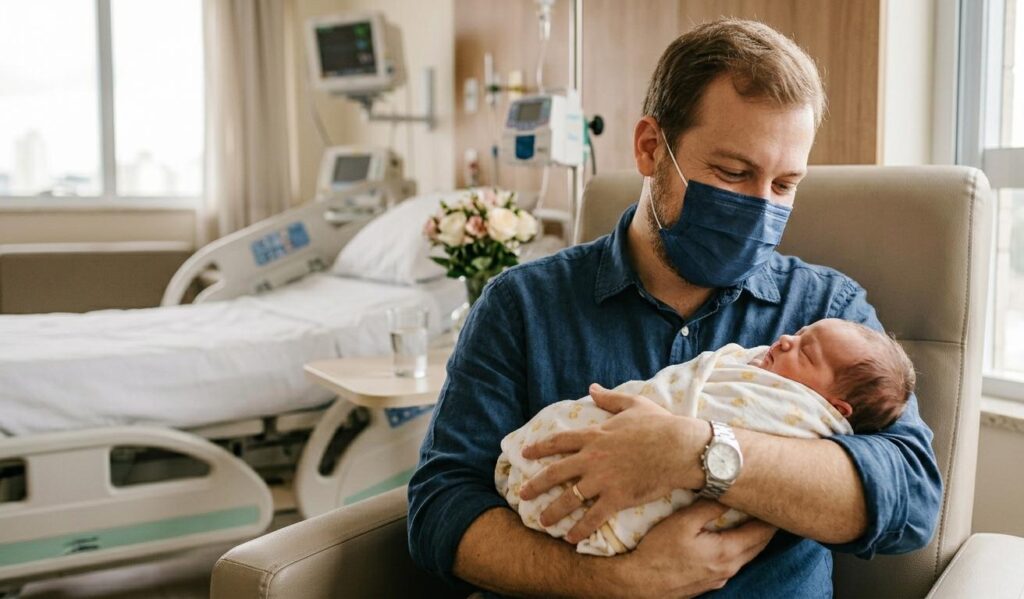 Homem vestindo camisa azul e máscara cirúrgica azul sentada em uma poltrona de hospital, segurando sorridente um bebê recém-nascido enrolado em uma manta branca com detalhes amarelos. Ao fundo, aparece um leito de maternidade, aparelhos médicos e um vaso de flores.