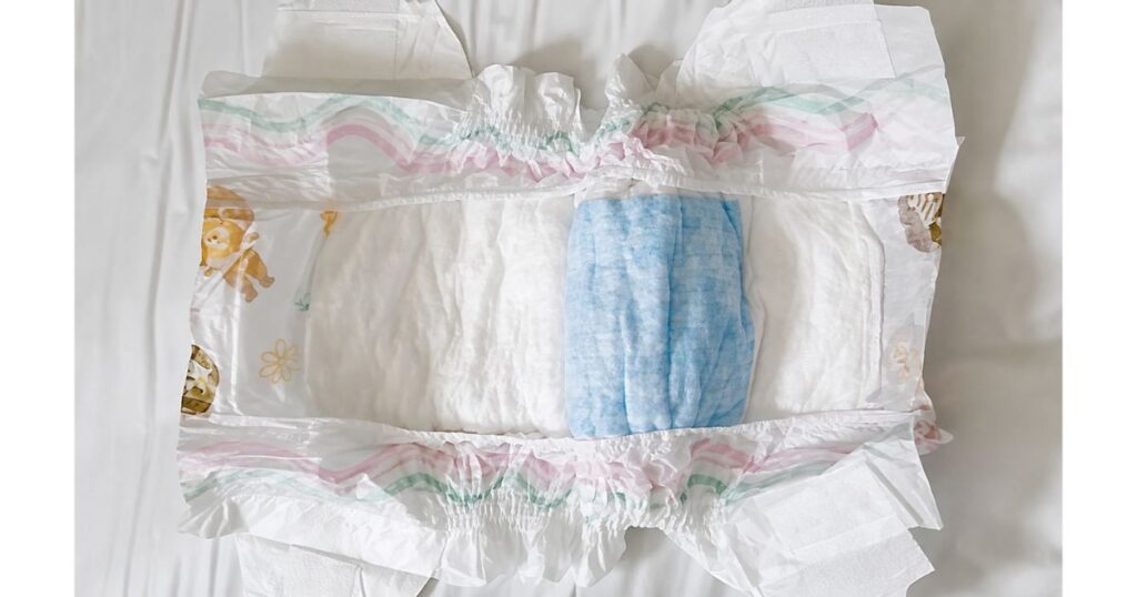 Fralda descartável infantil aberta por dentro, exibindo o núcleo de absorção branco com uma manta azul centralizada e elásticos nas bordas sobre superfície clara.