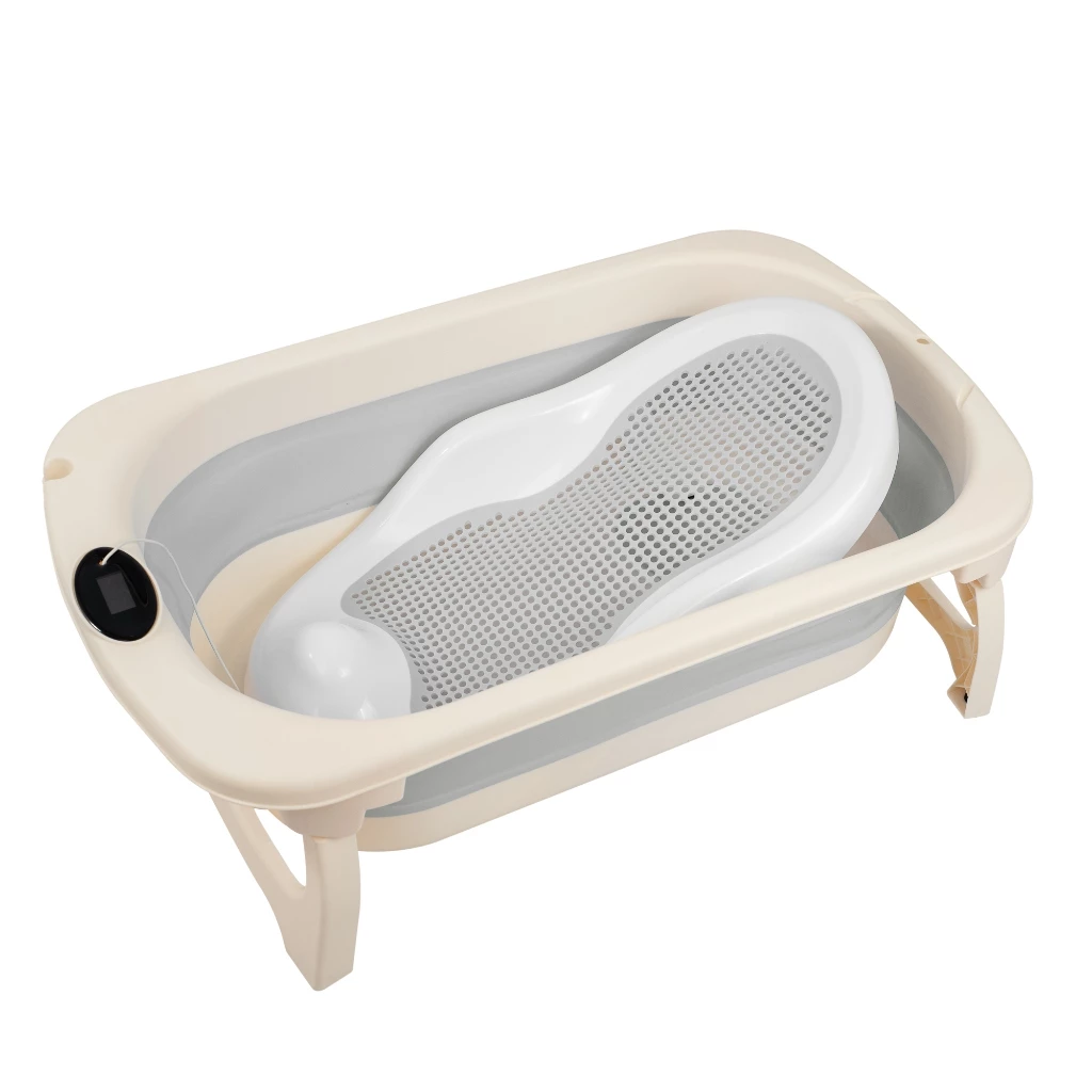 Banheira de bebê dobrável na cor bege e cinza com pernas de apoio abertas. No interior da banheira, há um assento redutor ergonômico branco com uma superfície cinza perfurada para apoiar o recém-nascido. Na borda esquerda da banheira, destaca-se um pequeno visor digital de termômetro redondo e preto. O fundo da imagem é totalmente branco.