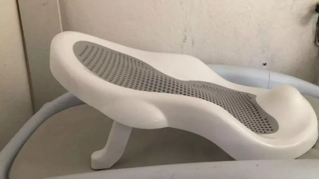 Assento redutor ergonômico para banheira de bebê, visto de perfil. O suporte possui estrutura plástica na cor branca com formato anatômico ondulado e uma área de encosto cinza com pequenos furos. Ele está apoiado por uma perna plástica branca inclinada sobre a base de uma banheira. O fundo mostra uma parede clara e texturizada.