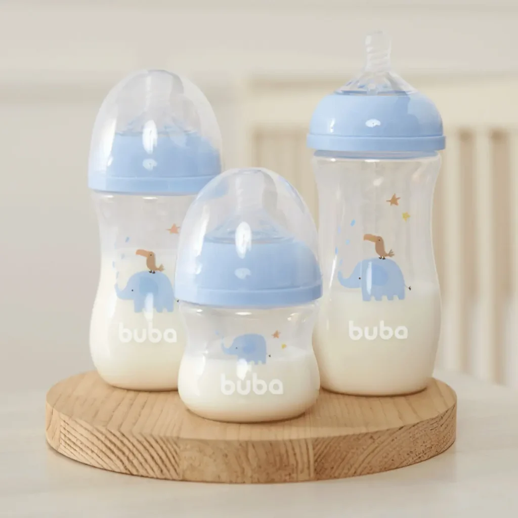 Três mamadeiras translúcidas da linha Buba Easy Flow com acabamento em azul-claro, contendo leite e organizadas por tamanho sobre uma base de madeira redonda. Os frascos possuem estampas sutis de elefante e pássaro em um cenário de quarto infantil com fundo claro e desfocado.
