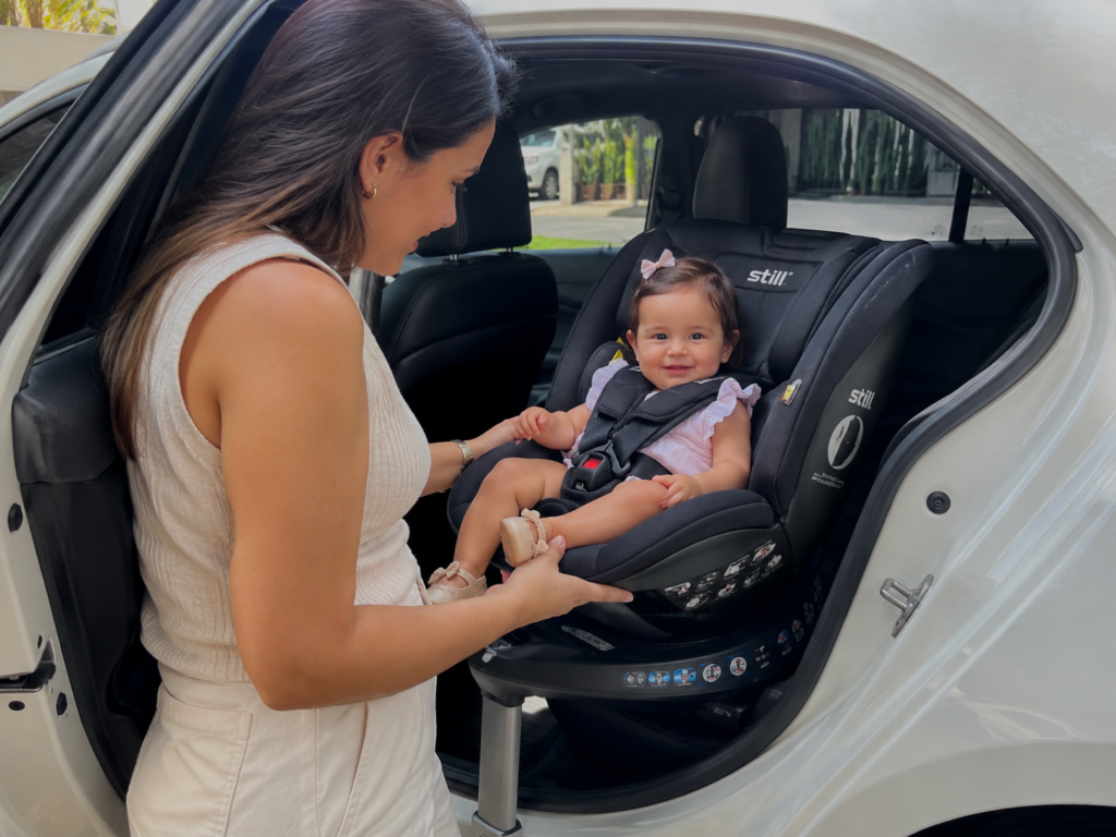 Mãe sorridente ao lado da porta aberta de um carro branco, interagindo com sua bebê que está sentada confortavelmente em uma cadeirinha preta da marca Stillo. A cadeirinha está rotacionada em direção à porta do veículo, demonstrando o sistema de giro 360 graus e o uso da haste de suporte frontal.