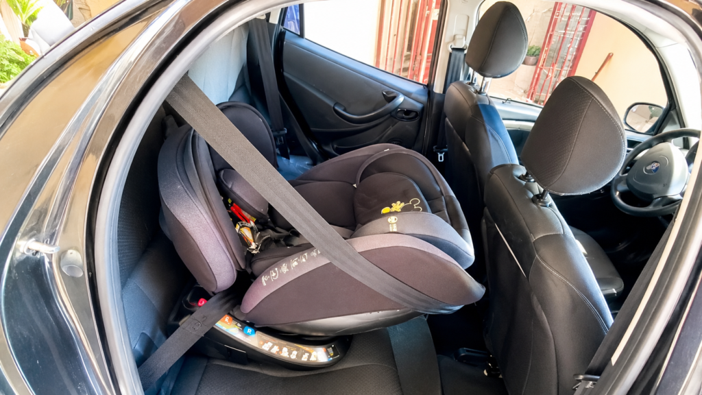 Vista lateral de uma cadeirinha de bebê preta com sistema de giro 360 graus instalada no banco traseiro de um carro com acabamento interno escuro. A cadeirinha está levemente rotacionada em direção à porta traseira aberta do veículo, demonstrando a facilidade de acesso.